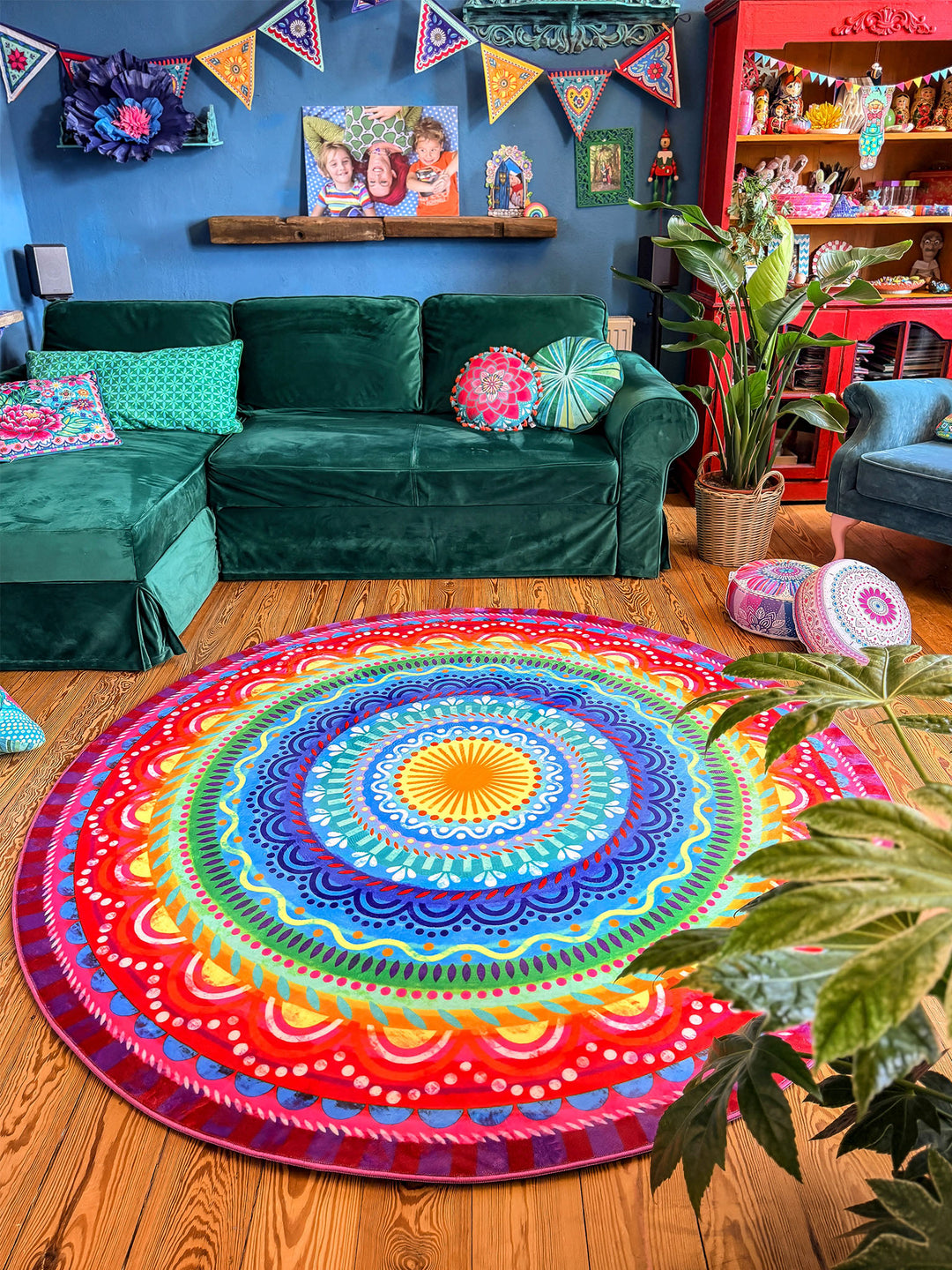 Runder Teppich mit farbenfrohem Mandala-Muster in Blau, Türkis, Gelb, Grün, Pink, Rot und Lila liegt auf warmem Holzboden in einem gemütlichen Wohnzimmer. Im Hintergrund stehen ein großes grünes Samtsofa, Zimmerpflanzen, bunte Kissen und dekorative Möbel vor einer dunkelblauen Wand mit Girlanden und Wandkunst. Der farbige Teppich setzt einen lebendigen Mittelpunkt und schafft eine kreative, wohnliche Boho-Atmosphäre