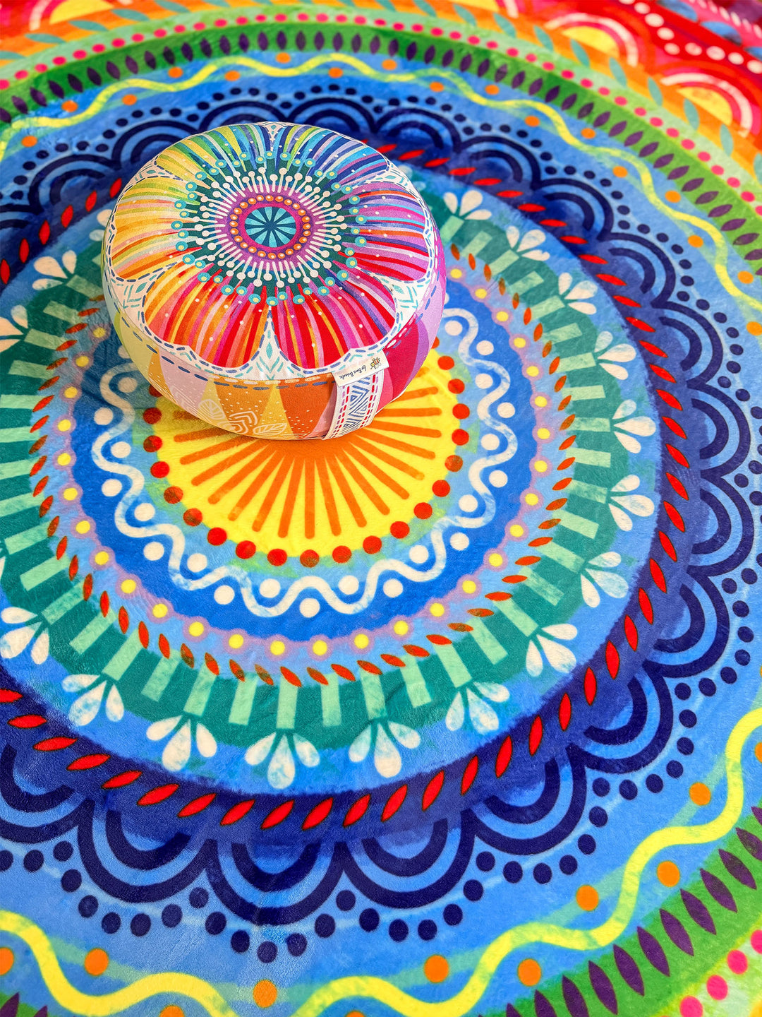 Buntes Yogakissen mit floralem Muster in Pink, Gelb, Orange, Blau und Türkis steht mittig auf einem runden Teppich mit farbenfrohem Mandala-Design. Der Teppich zeigt kreisförmige Ornamente in Blau, Türkis, Grün, Gelb, Rot und Orange. Die Nahaufnahme betont die leuchtenden Farben, detailreichen Muster und die gemütliche, kreative Wohnatmosphäre im Boho-Stil.