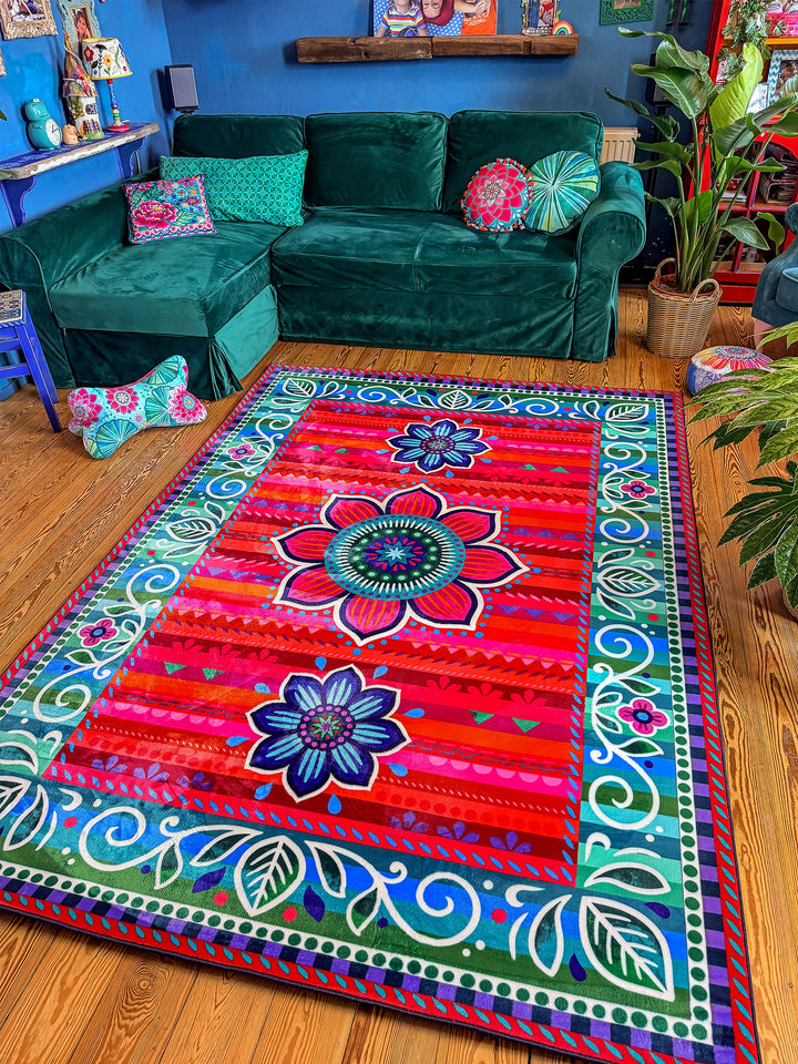 Großer rechteckiger Teppich mit farbenfrohem Blumen- und Ornamentmuster in Rot, Pink, Türkis, Blau, Grün und Lila liegt auf warmem Holzboden in einem gemütlichen Wohnzimmer. Im Hintergrund stehen ein grünes Samtsofa, ein blauer Sessel, Zimmerpflanzen und dekorative Möbel vor einer dunkelblauen Wand mit bunten Girlanden. Der bunte Teppich setzt einen lebendigen Mittelpunkt und schafft eine kreative, wohnliche Boho-Atmosphäre.