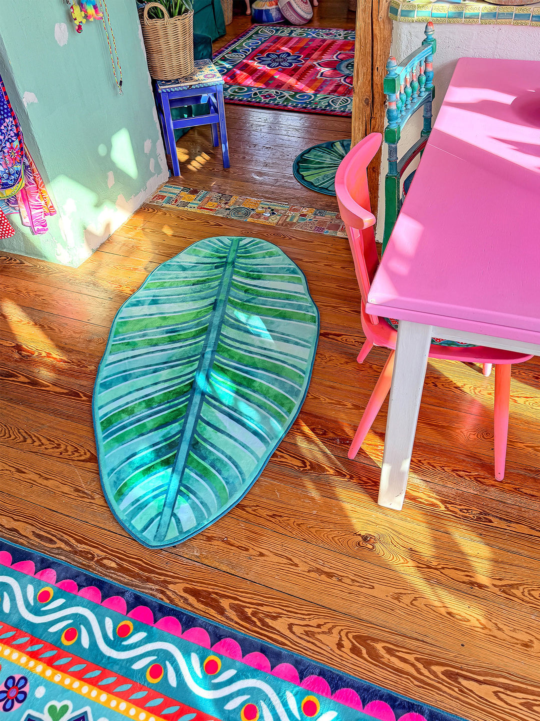 Grüner Blatt-Teppich liegt auf warmem Holzboden in einem farbenfrohen Wohnraum mit kreativer Einrichtung. Im Hintergrund stehen viele Zimmerpflanzen, ein pinker Tisch mit passenden Stühlen sowie weitere bunte Teppiche mit Mandala- und Blumenmustern. Sonnenlicht fällt in den Raum und betont die natürlichen Farben. Die Szene wirkt gemütlich, verspielt und tropisch inspiriert.