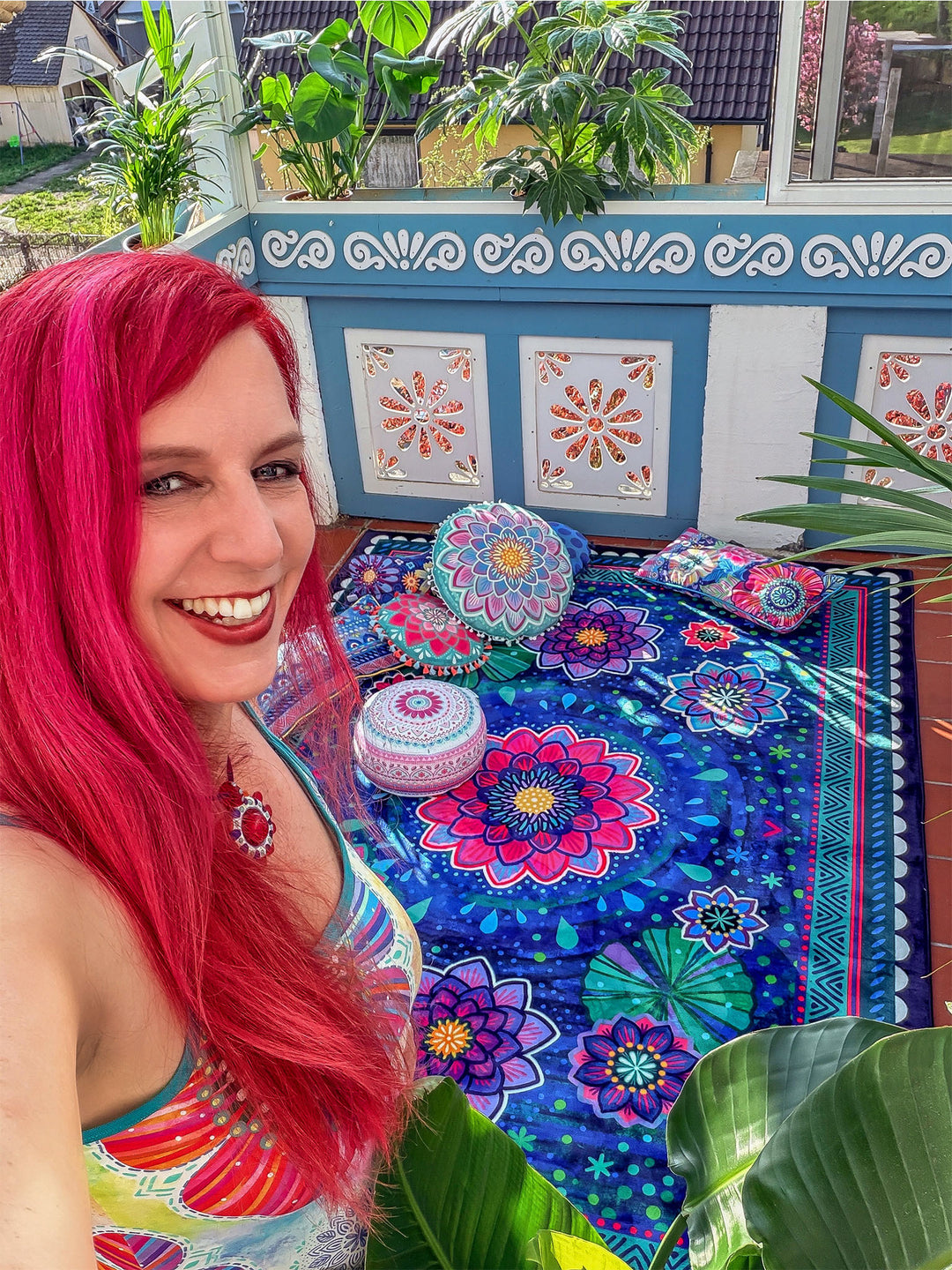 Bine Brändle lächelt in die Kamera auf einem hellen Wintergarten oder Balkon mit vielen grünen Pflanzen. Im Hintergrund liegt ein großer Teppich mit floralem Mandala-Muster in Dunkelblau, Pink, Lila, Türkis und Grün, dekoriert mit bunten Kissen und Sitzhockern. Weiße und blaue Wanddetails sowie Sonnenlicht schaffen eine freundliche, gemütliche Wohnatmosphäre mit kreativem Boho-Stil.