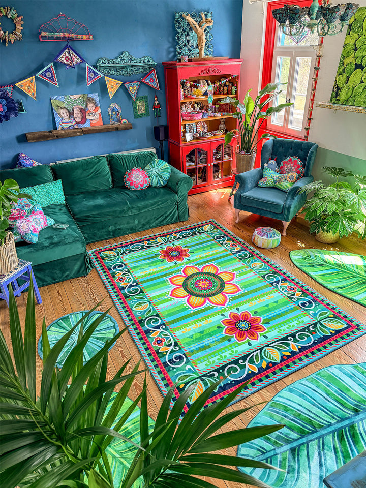 Stilvolles Wohnzimmer im farbenfrohen Boho-Stil mit großem rechteckigem Teppich in Grün, Türkis und Pink mit Blumenmandala-Muster in der Mitte. Rundherum liegen mehrere blattförmige Teppiche und runde Blattmatten in verschiedenen Grüntönen auf warmem Holzboden. Eine dunkelgrüne Samt-Couch, ein petrolfarbener Sessel, viele Zimmerpflanzen sowie ein leuchtend roter Vitrinenschrank setzen starke Farbakzente. Die dunkelblaue Wand mit Wimpelkette, Bildern und dekorativen Details schafft eine kreative, gemütliche 