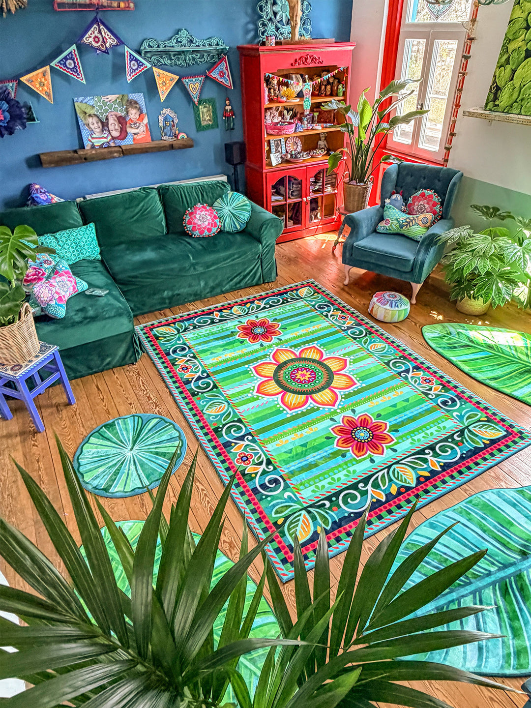 Stilvolles Wohnzimmer im farbenfrohen Boho-Stil mit großem rechteckigem Teppich in Grün, Türkis und Pink mit Blumenmandala-Muster in der Mitte. Rundherum liegen mehrere blattförmige Teppiche und runde Blattmatten in verschiedenen Grüntönen auf warmem Holzboden. Eine dunkelgrüne Samt-Couch, ein petrolfarbener Sessel, viele Zimmerpflanzen sowie ein leuchtend roter Vitrinenschrank setzen starke Farbakzente. Die dunkelblaue Wand mit Wimpelkette, Bildern und dekorativen Details schafft eine kreative, gemütliche 