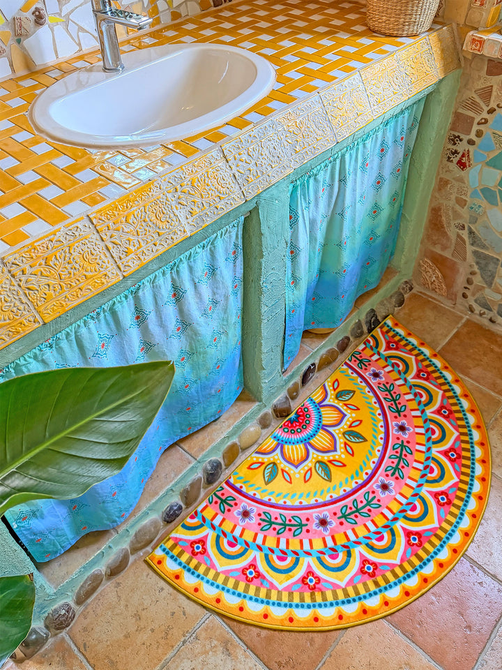 Halbrunder Badteppich in leuchtendem Gelb mit farbenfrohem Mandala- und Blumenmuster in Pink, Türkis, Orange, Blau und Grün liegt auf warmen Natursteinfliesen vor einem Waschtisch. Der rustikal-kreative Waschtisch besitzt eine gelb-weiß geflieste Ablage, ein weißes Waschbecken und türkisfarbene Stoffvorhänge darunter. Naturstein- und Mosaikdetails an Wand und Möbeln verleihen dem Badezimmer einen kunstvollen Boho-Stil. Die fröhliche Matte setzt einen lebendigen Farbakzent im Raum.