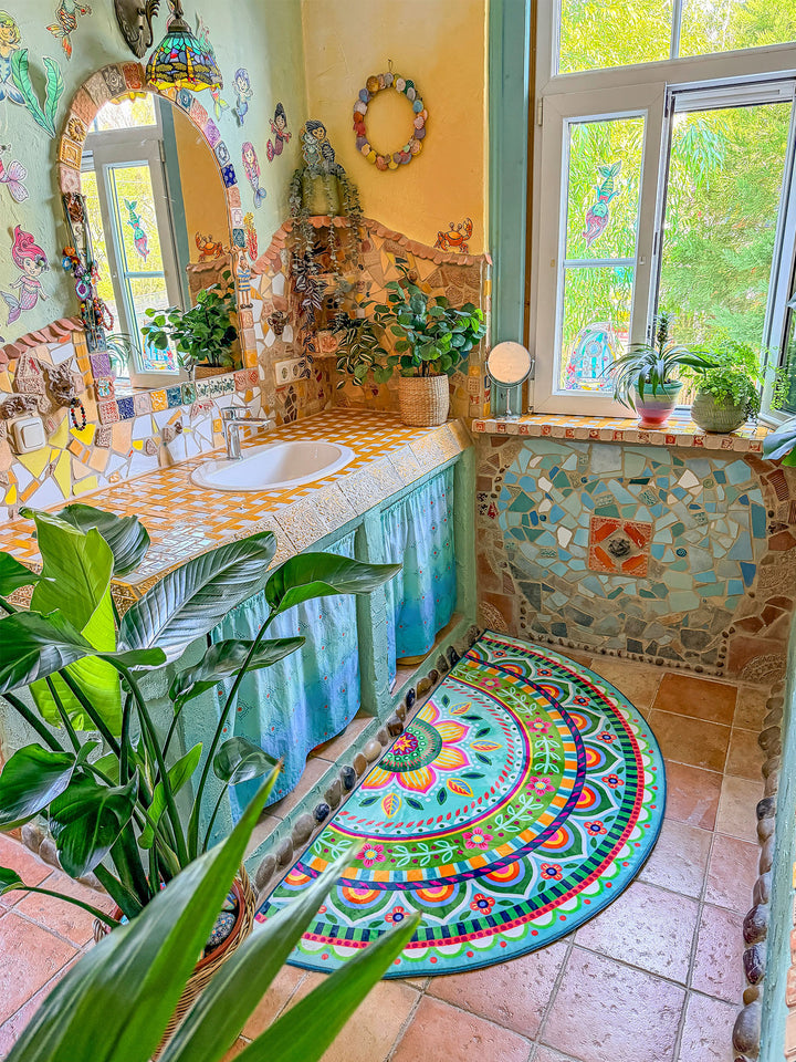 Halbrunder Badteppich in frischen Türkis-, Grün-, Pink- und Gelbtönen liegt auf beigefarbenen Fliesen vor einem kunstvoll gestalteten Waschbecken im Boho-Stil. Das Teppichdesign zeigt ein großes Mandala mit floralen Ornamenten, Blütenranken und bunten Bordüren. Darüber befindet sich ein Waschtisch mit gelb-weißer Mosaikplatte, integriertem Waschbecken und türkisfarbenen Stoffvorhängen als Unterschrank. Die Wände sind mit Naturstein, bunten Fliesen, Spiegelbogen und verspielten Meerjungfrauen-Motiven dekorie