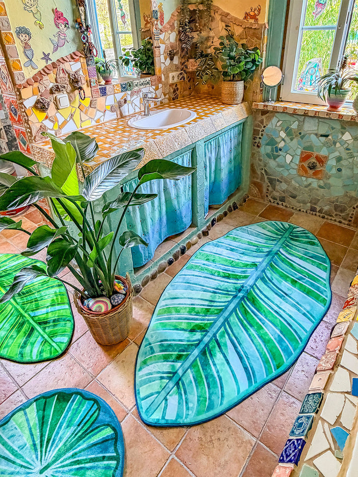 Künstlerisch gestaltetes Badezimmer im Boho- und Tropenstil mit mehreren grünen Badteppichen in Blattform auf terrakottafarbenen Fliesen. Die Teppiche erinnern an große tropische Blätter mit detailreichen Blattadern in verschiedenen Grün- und Türkistönen. Im Mittelpunkt steht eine große Zimmerpflanze im Korb, umgeben von den Teppichen. Der Waschtisch ist mit gelb-weißen Mosaikfliesen verkleidet und besitzt türkisfarbene Stoffvorhänge als Stauraum. Wände, Fensterrahmen und Badewanne sind mit Naturstein, bunt