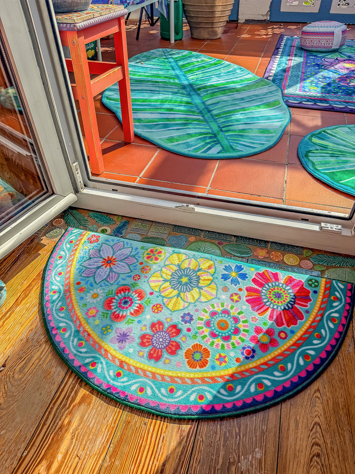 Halbrunder Teppich in Türkis liegt vor einer geöffneten Terrassentür auf warmem Holzboden. Der Teppich ist mit bunten Blumen, Mandala-Ornamenten und verspielten Mustern in Pink, Gelb, Rot, Orange, Blau und Grün gestaltet. Im Außenbereich auf roten Fliesen liegen grüne Blatt-Teppiche. Die helle Szene verbindet farbenfrohes Wohndesign mit natürlicher, sommerlicher Wohnatmosphäre.